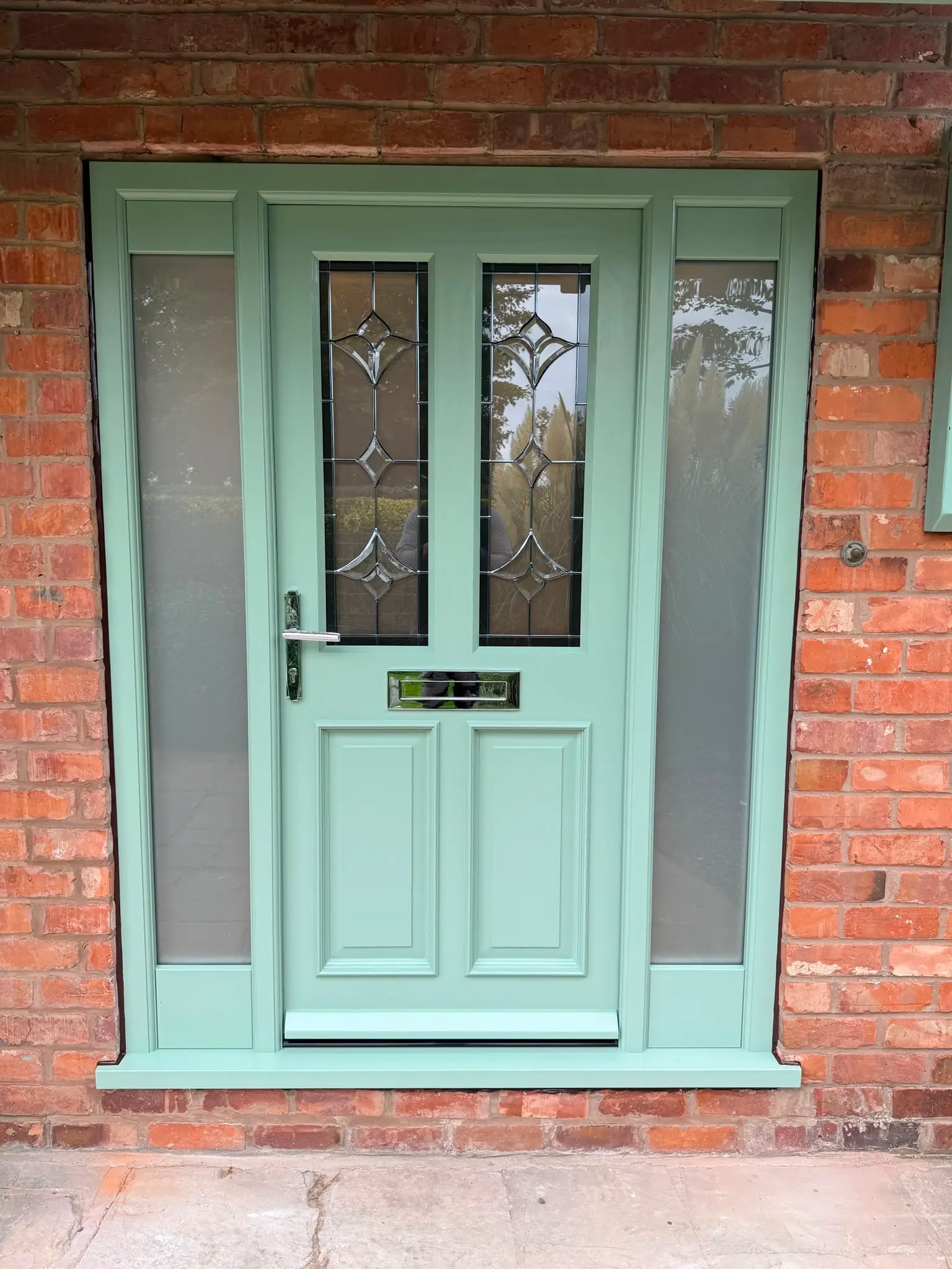 Sage green front door with bevelled leaded glazing and matching glazed sidelights, with chrome furniture and letterplate, set into red brick.