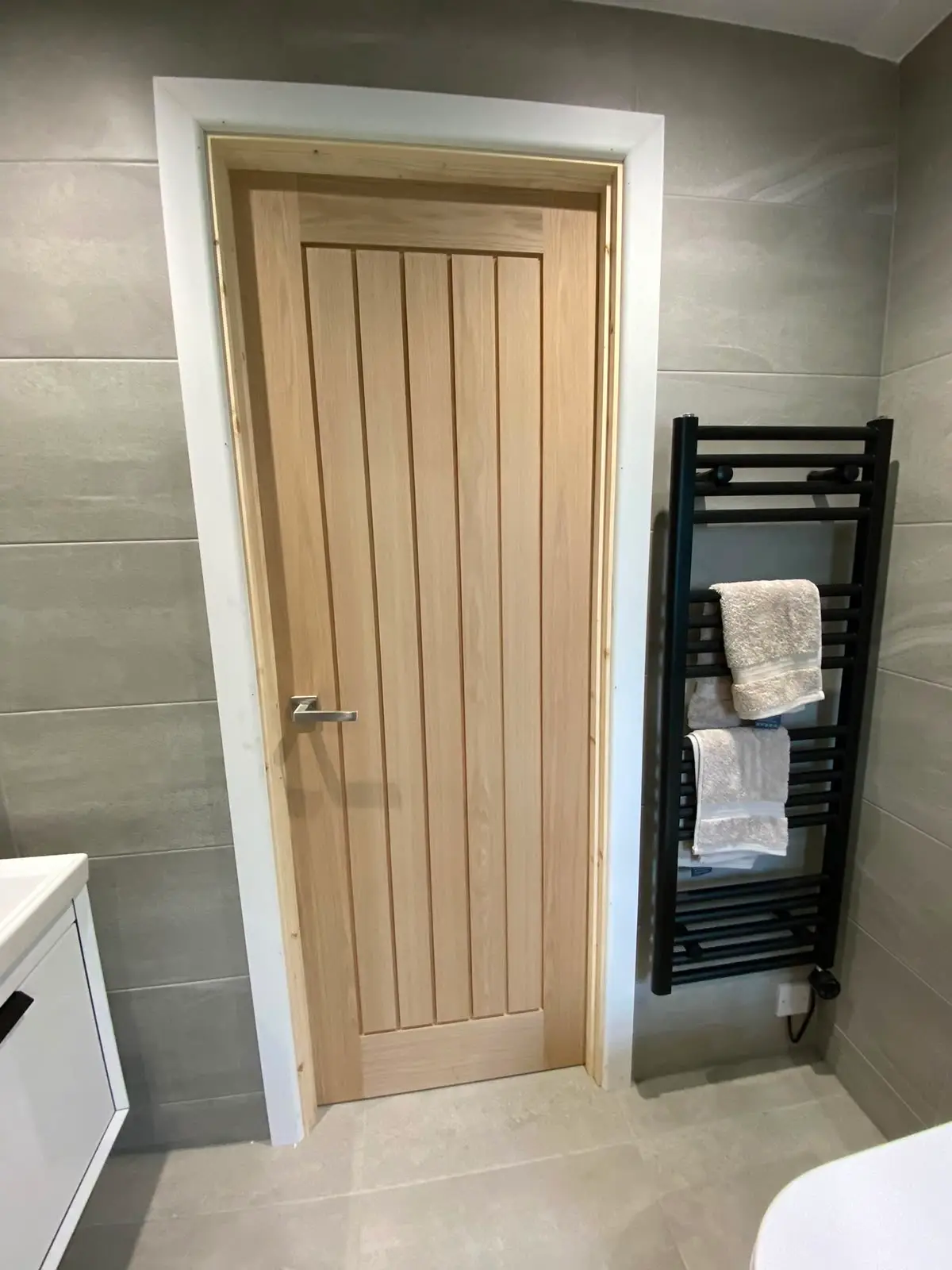 Oak vertical-boarded door fitted to a tiled bathroom, hung in a bare timber lining with a chrome lever handle.