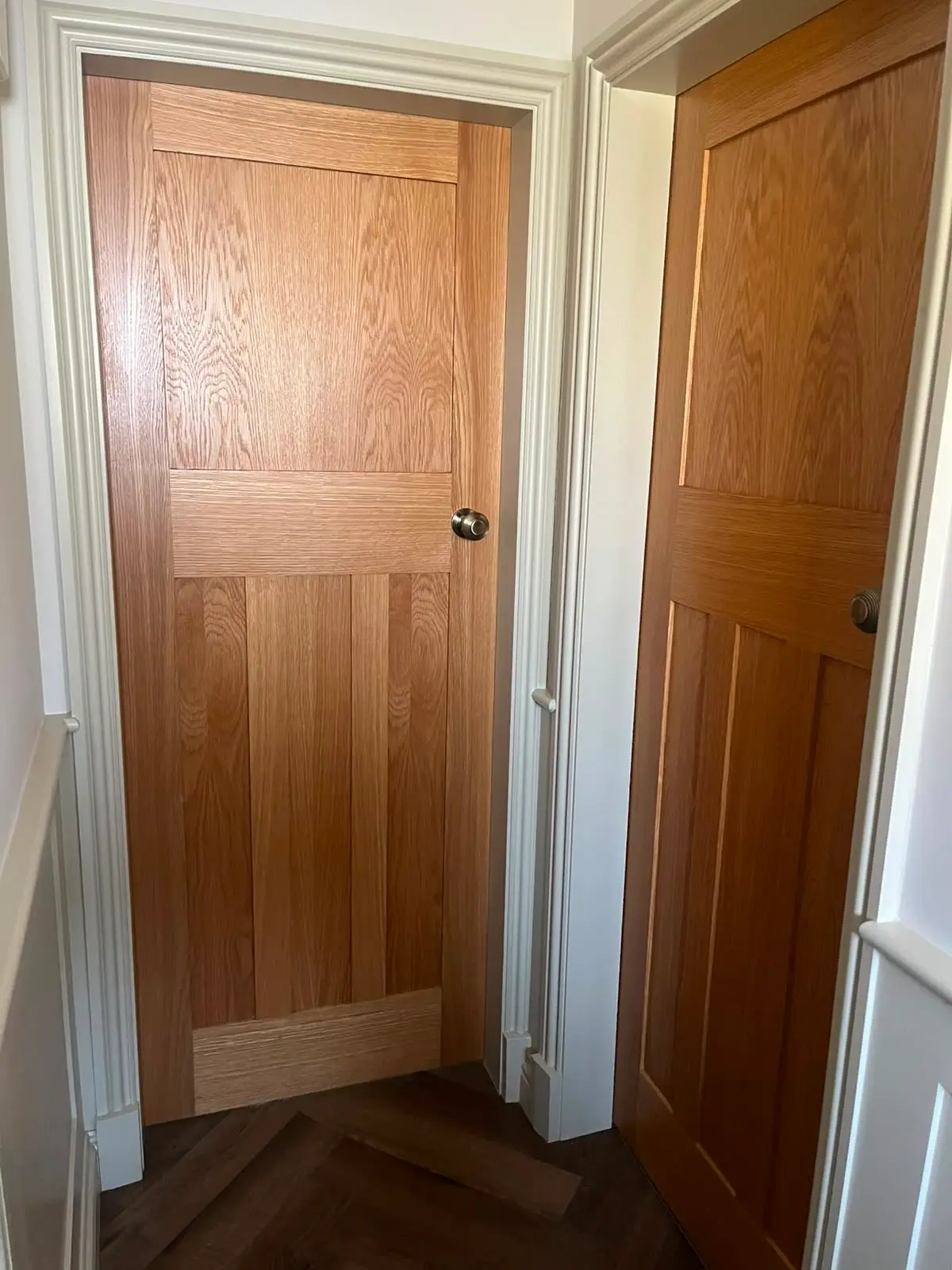 Two oak four-panel doors hung in adjacent white painted linings, opening onto a herringbone parquet floor, with brushed knobs and moulded architrave.