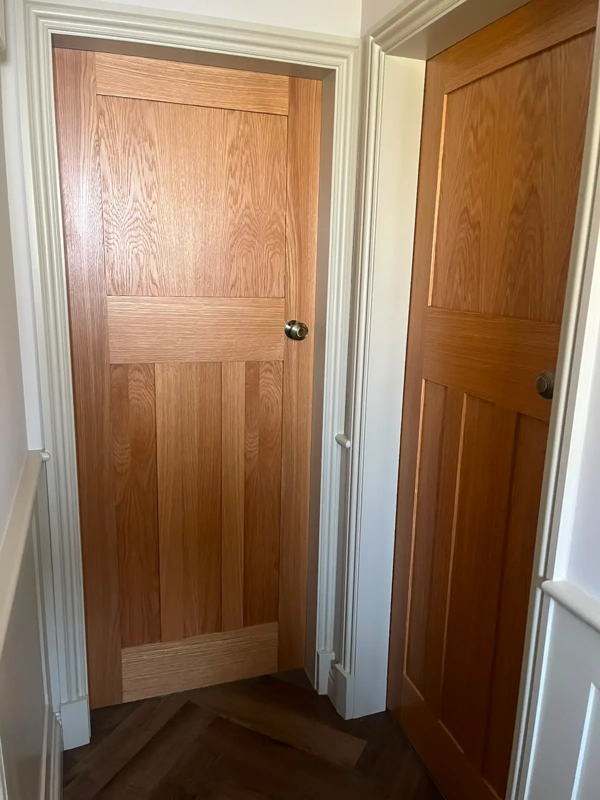 Two oak four-panel doors hung in adjacent white painted linings, opening onto a herringbone parquet floor, with brushed knobs and moulded architrave.