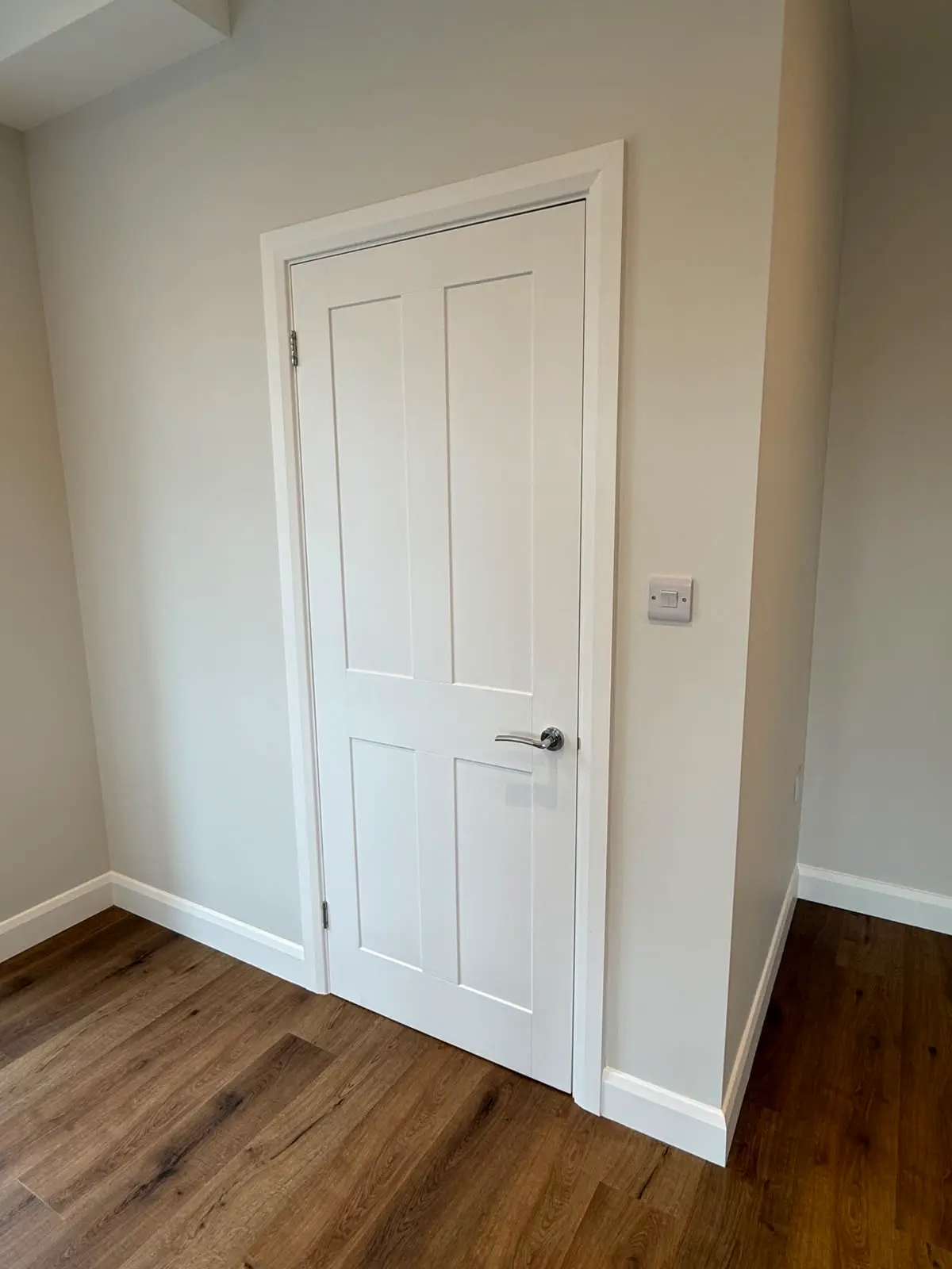 White painted four-panel door hung in a new lining with moulded architrave and a chrome lever handle, against an oak floor.