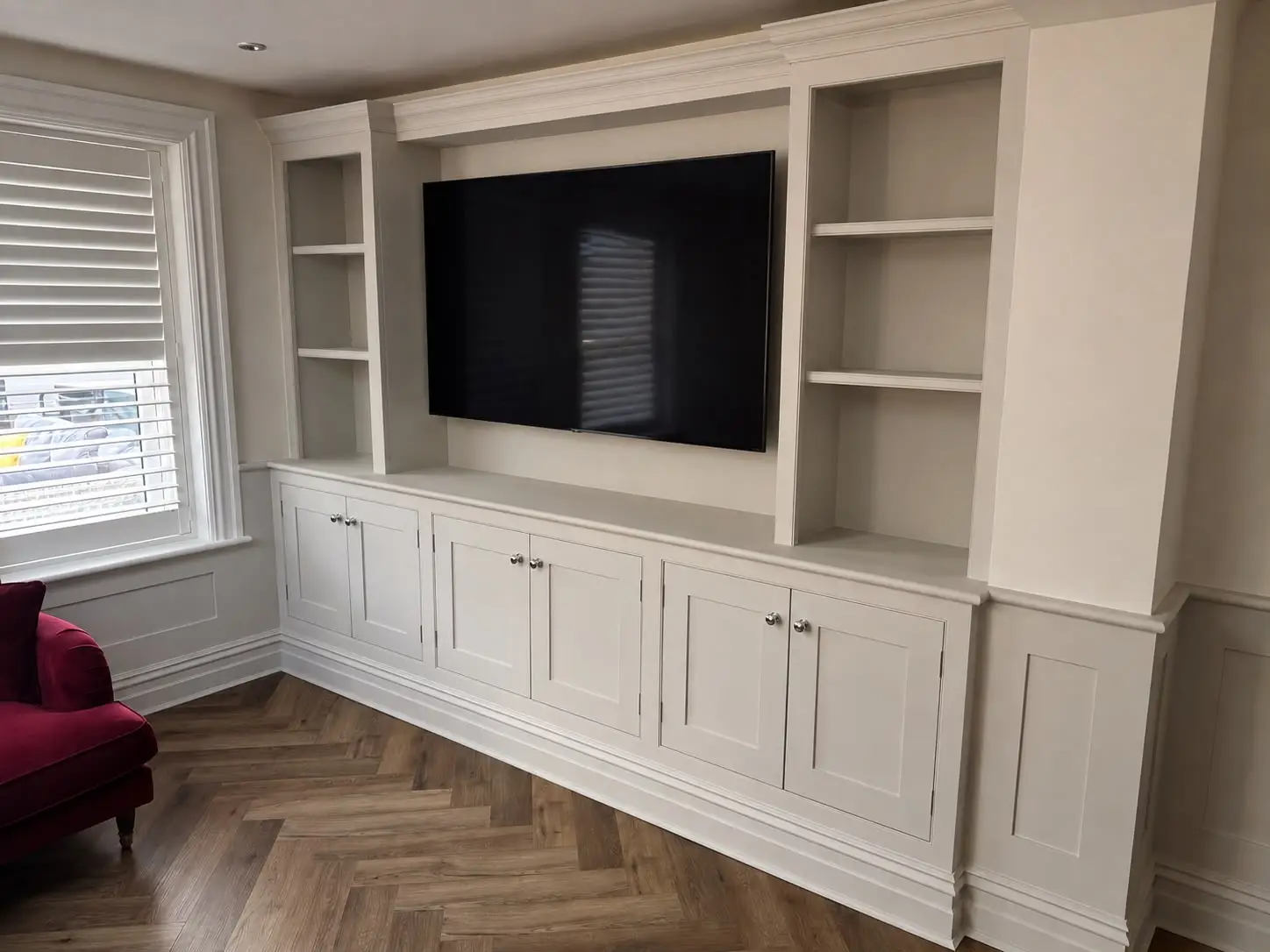Fitted media wall in off-white shaker style, with open shelving either side of a recessed television space above a run of panelled cupboards, finished with a moulded cornice and skirting to match.