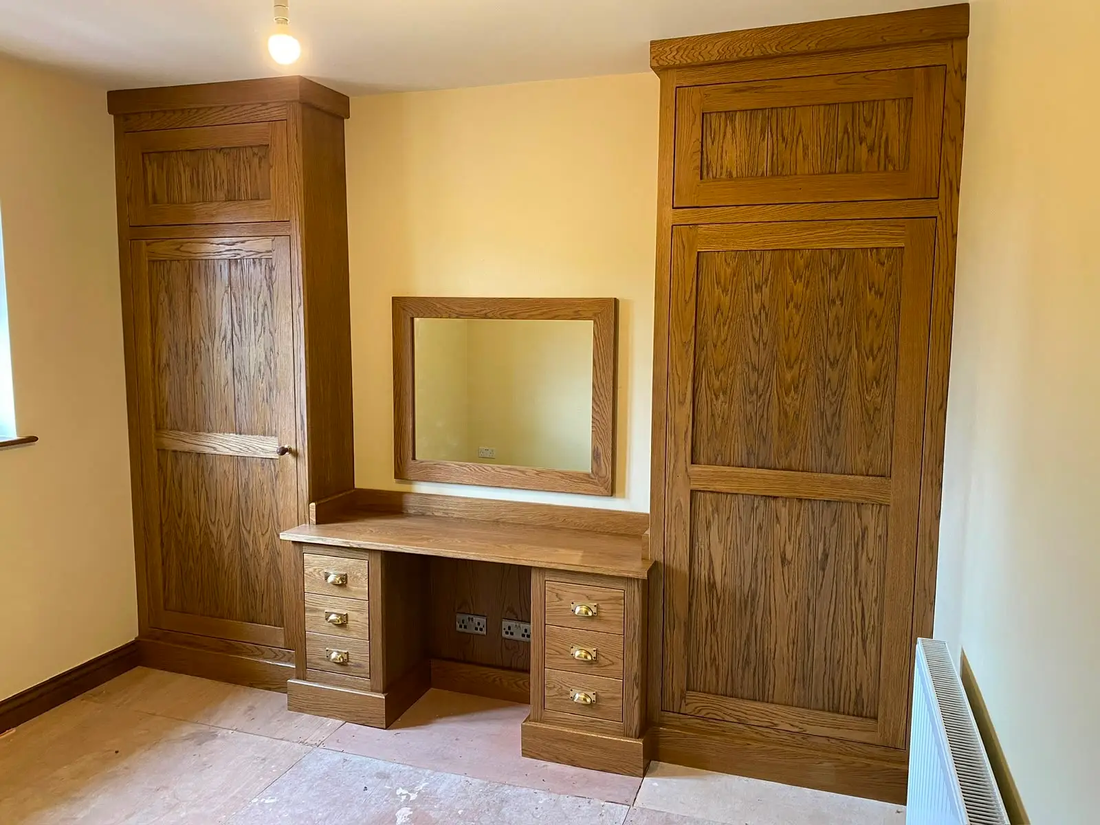 Fitted oak bedroom furniture: full-height panelled wardrobes flanking a dressing table with drawers and a matching oak mirror.