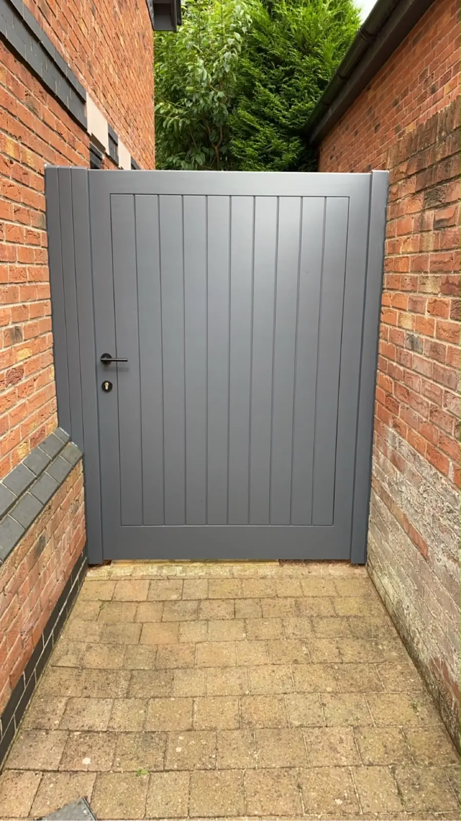 Grey painted timber side gate with vertical boarding, hung between brick walls on a block-paved path.