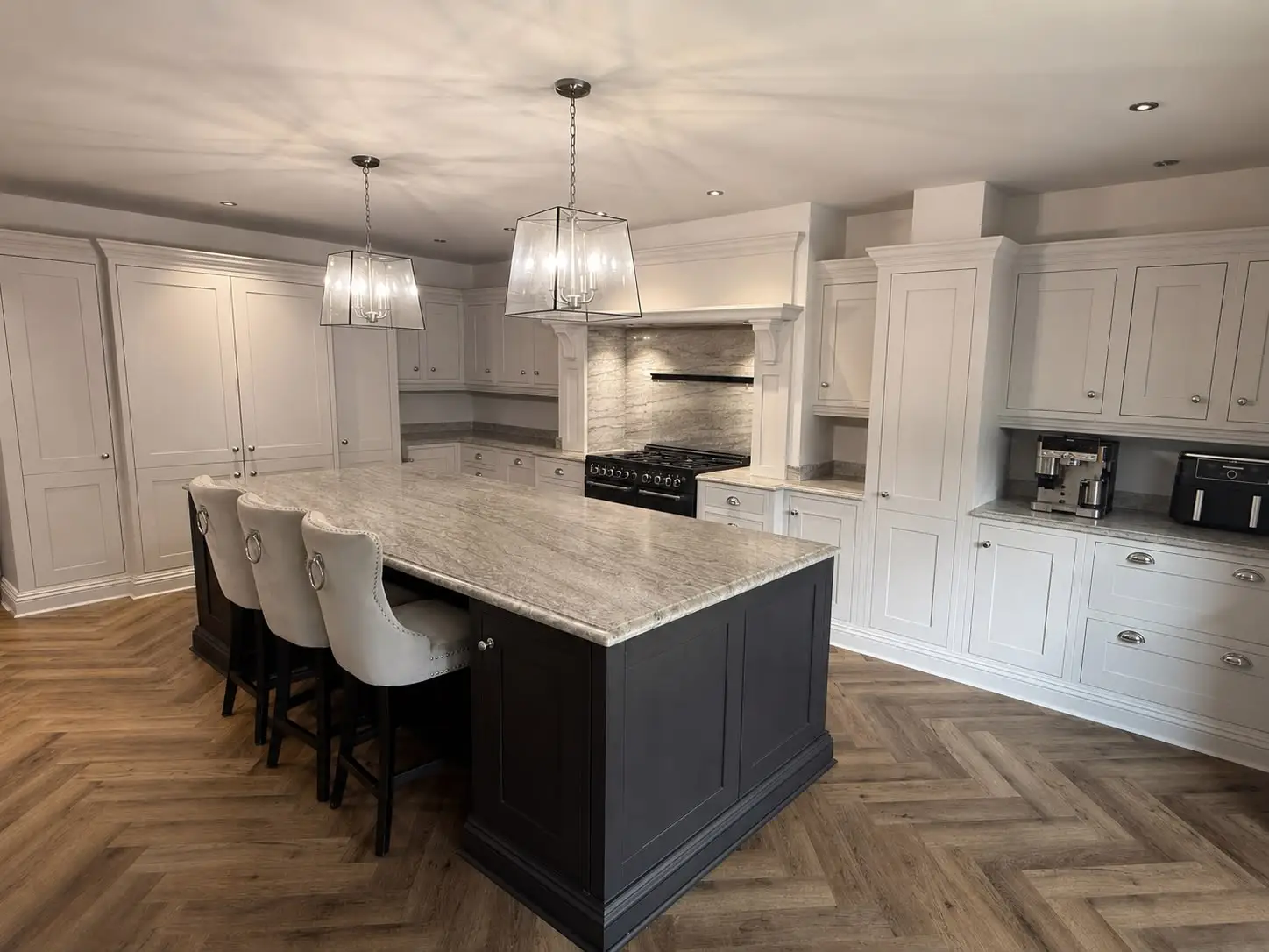Pale grey-white shaker kitchen with a contrasting dark island under a grey marble-effect worktop, matching upholstered bar stools and a black range cooker set into a marble-clad chimney breast, over a herringbone floor.