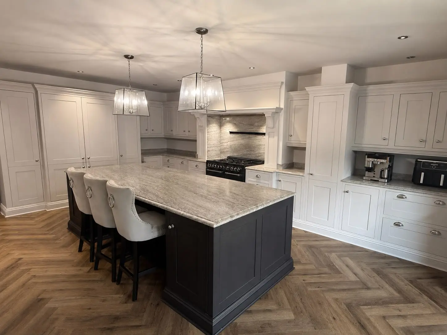 Pale grey-white shaker kitchen with a contrasting dark island under a grey marble-effect worktop, matching upholstered bar stools and a black range cooker set into a marble-clad chimney breast, over a herringbone floor.