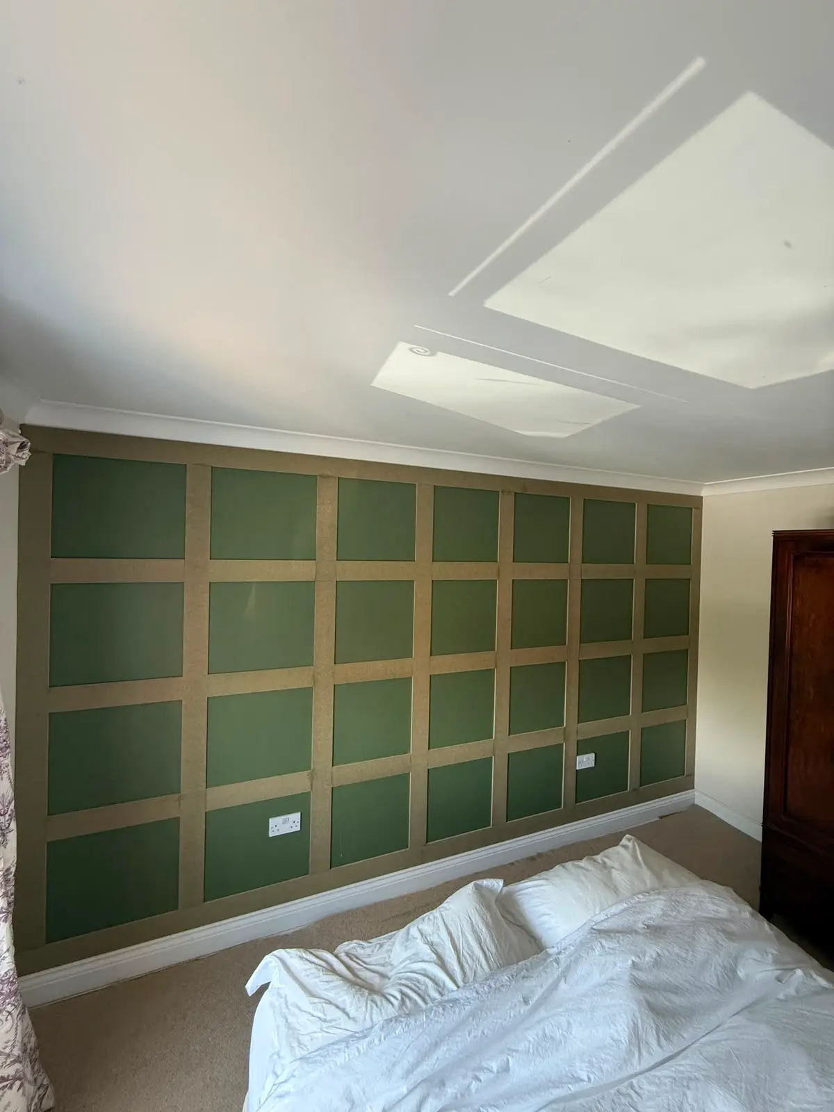 Wall panelling part-installed in a bedroom, bare board framework set out in a grid over dark green paint, before filling and painting.