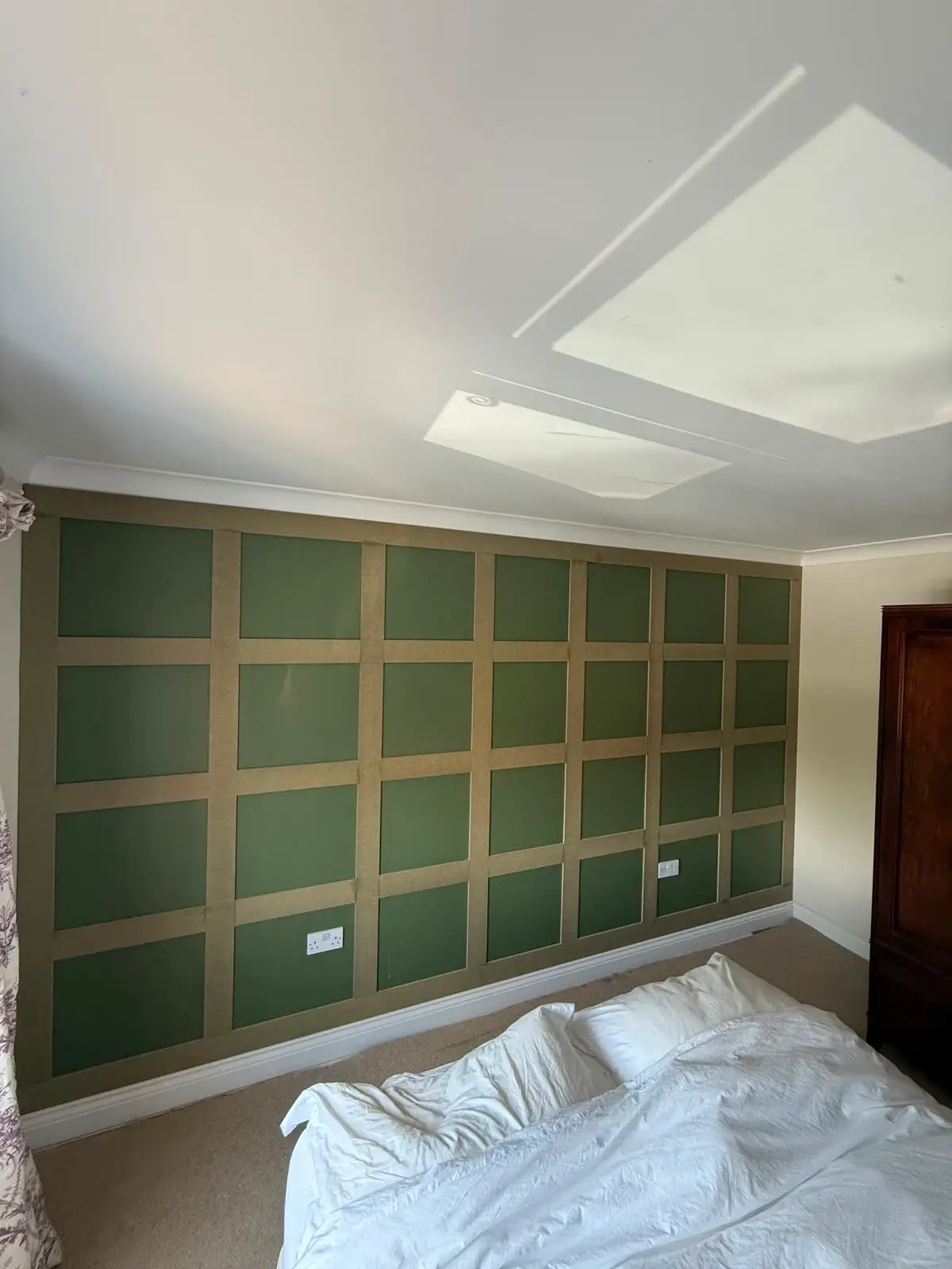 Wall panelling part-installed in a bedroom, bare board framework set out in a grid over dark green paint, before filling and painting.