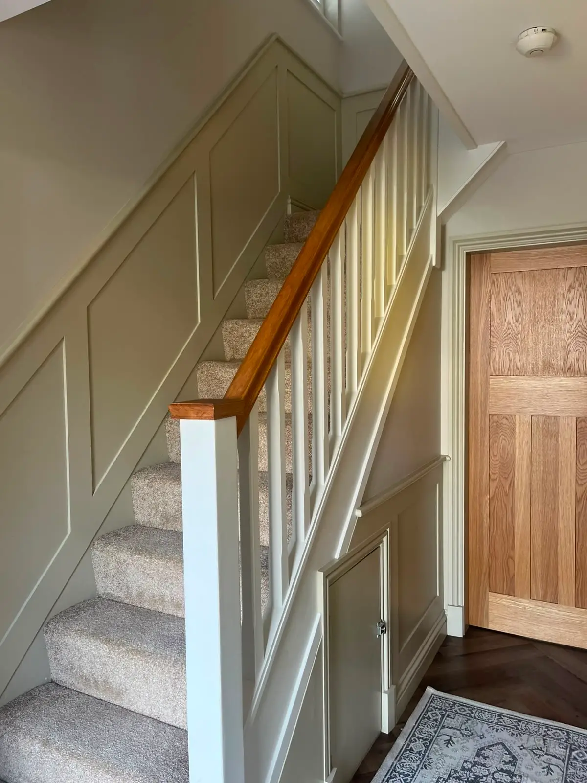 Sage panelling to a staircase wall and understair door, with white spindles, an oak handrail and an oak boarded door alongside.