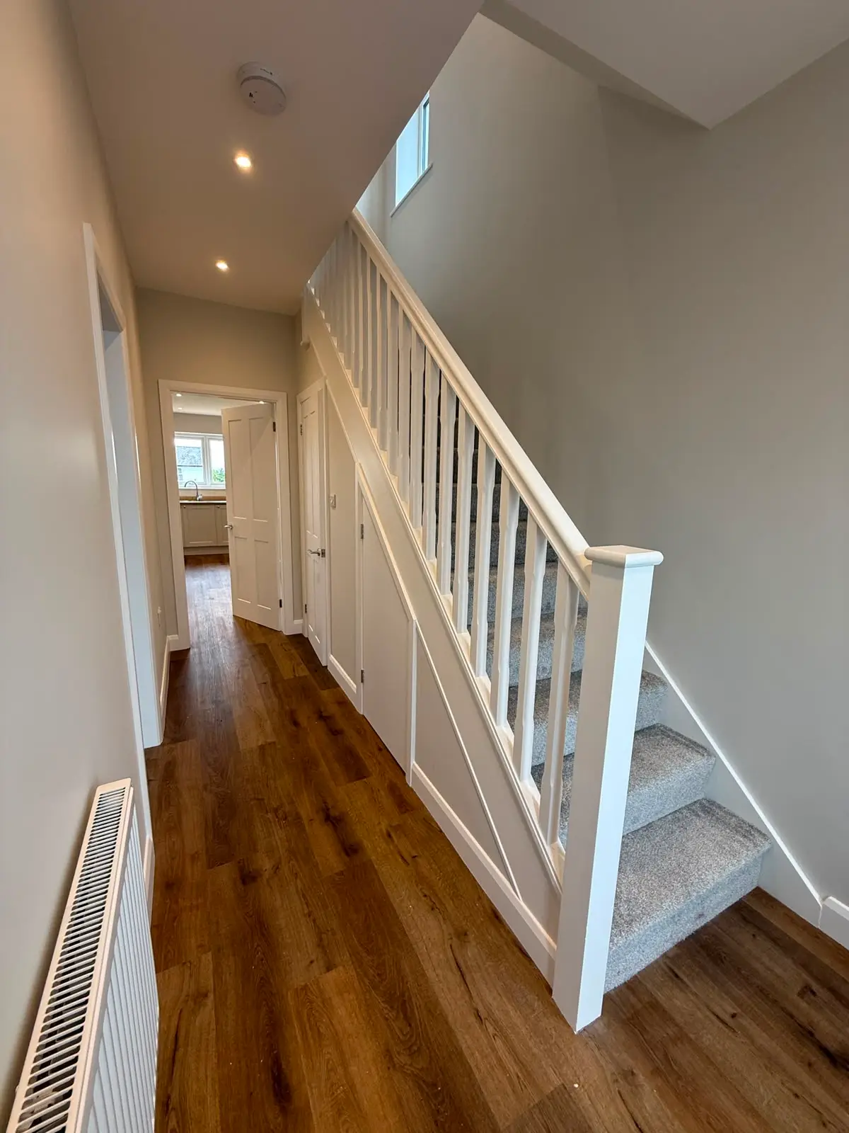 White painted staircase with turned spindles and a grey carpet runner, rising from a hallway with an oak-effect floor and panelled understair storage.
