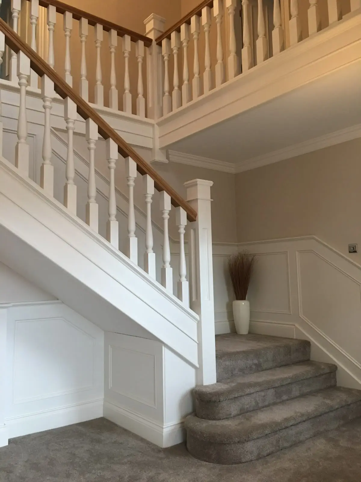 White wall panelling following the line of a staircase in a hallway, below turned spindles and an oak handrail.