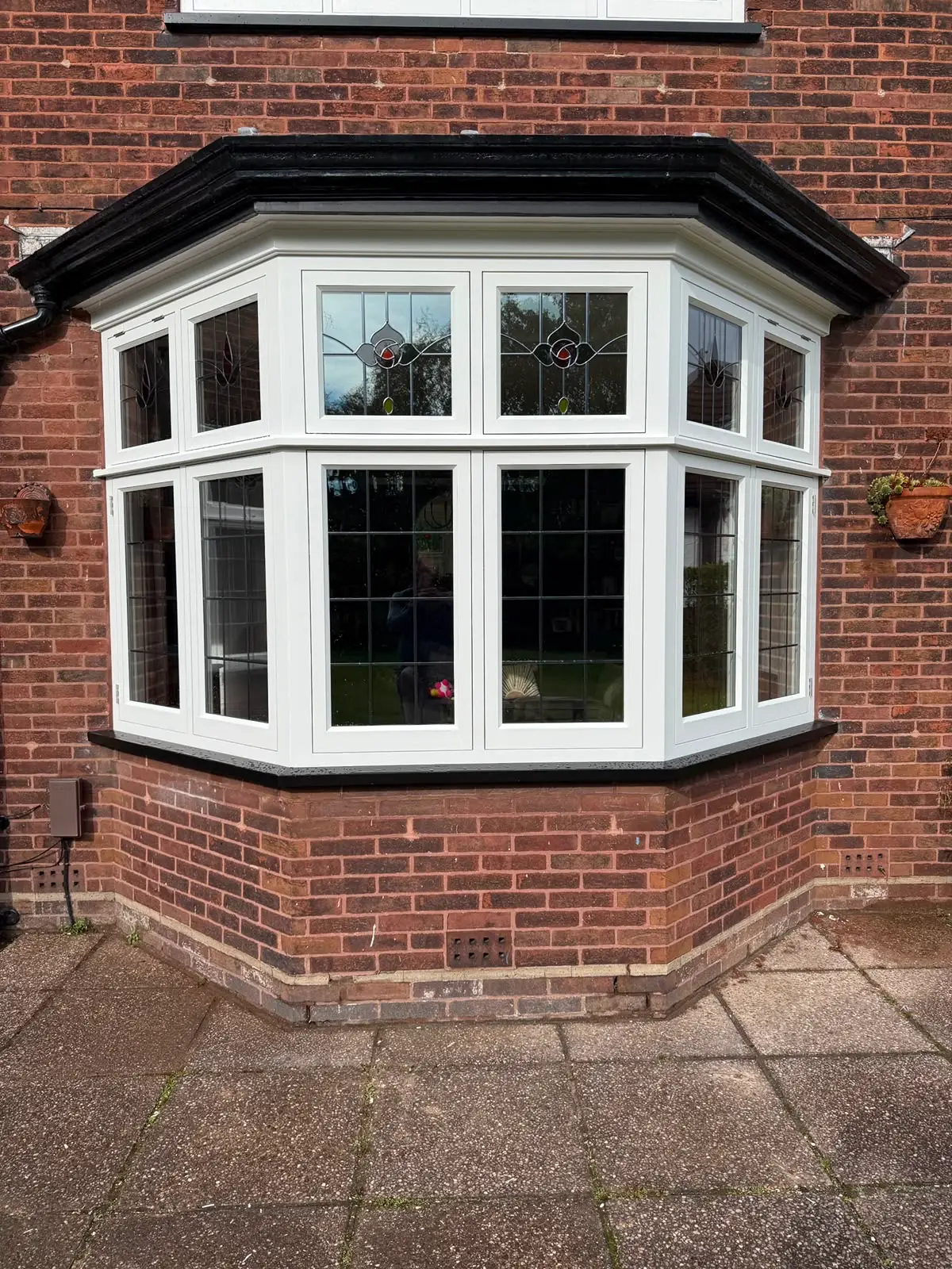 Splayed bay window in white painted timber, its upper lights glazed with leaded stained glass in a rose motif above square-leaded panes.